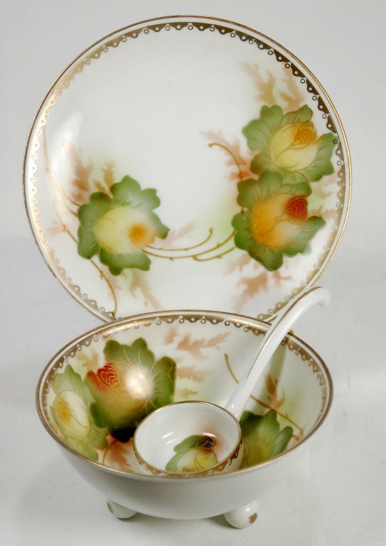 This lovely complete set of a condiment bowl, saucer and ladle features a Japanese influenced floral motif. The dainty bowl sits on three balled feet. The ladle has a curved handle and a small flower inside of the dipper. The saucer mimics the