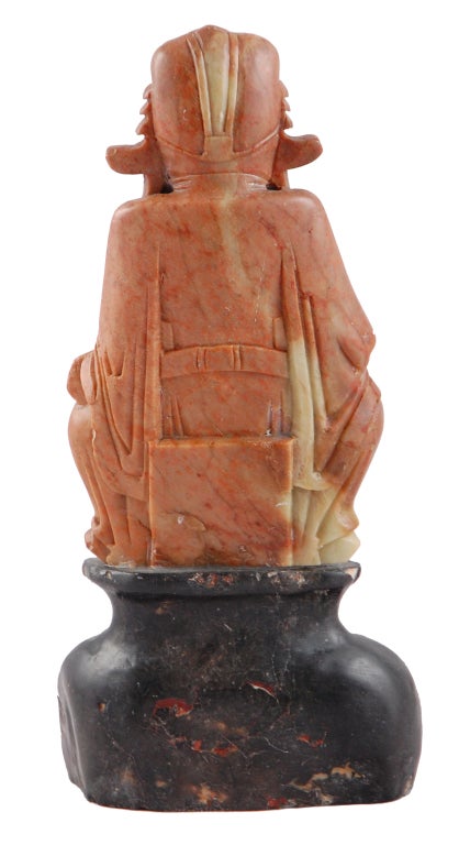 Cette figurine sculptée de style asiatique est faite de pierre de savon veinée de rose et de blanc. Il repose sur une base noire sculptée. Ce personnage assis tient un cadeau dans sa main et vous portera chance.
