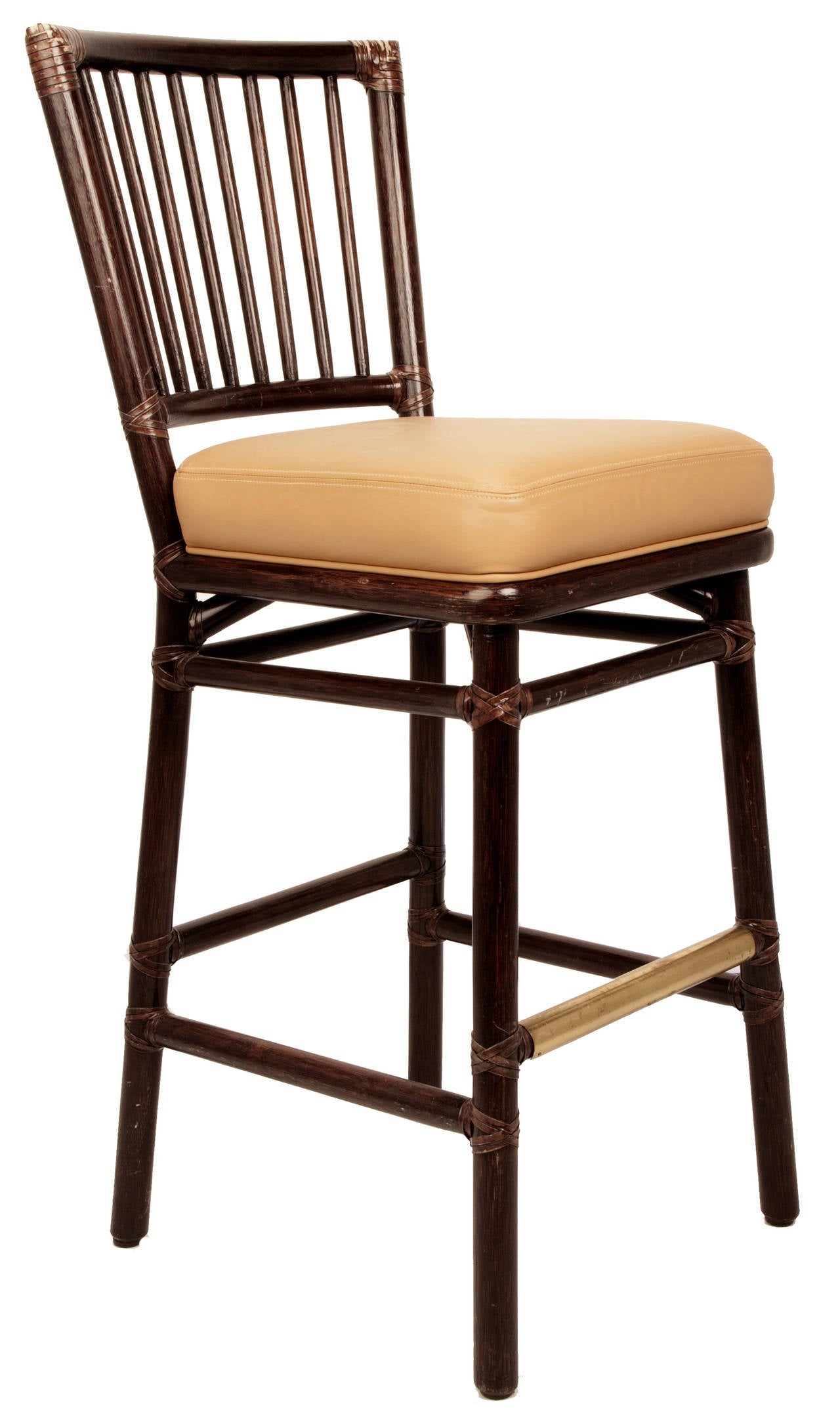 These McGuire bar stools with rattan frames and typical leather wrapped joinery, the backs have vertical spindles, the foot rail is covered in brass and the seats are upholstered in wheat colored leather with a self welt