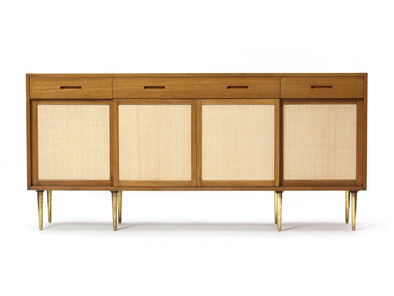 A credenza or cabinet with three drawers over four sliding linen-front doors with turned brass legs.