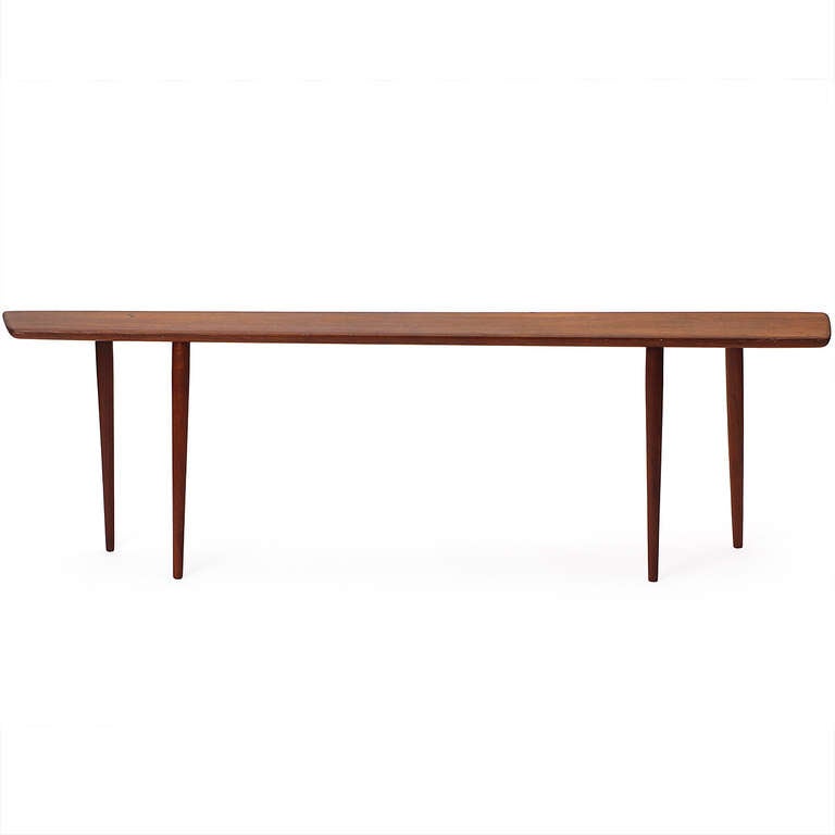 An unusual and expressive narrow occasional or display table in teak having a  trapezoidal single board top and four tall turned legs that can be attached in different configurations.