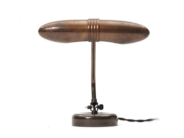 A patinated brass desk lamp with an articulated capsule-form shade and a bent, adjustable arm on a circular base.