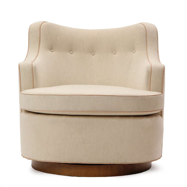 A graceful pair of swiveling lounge chairs of barrel-back form upholstered in a cream wool with natural leather welting, and resting on a walnut disc bases.