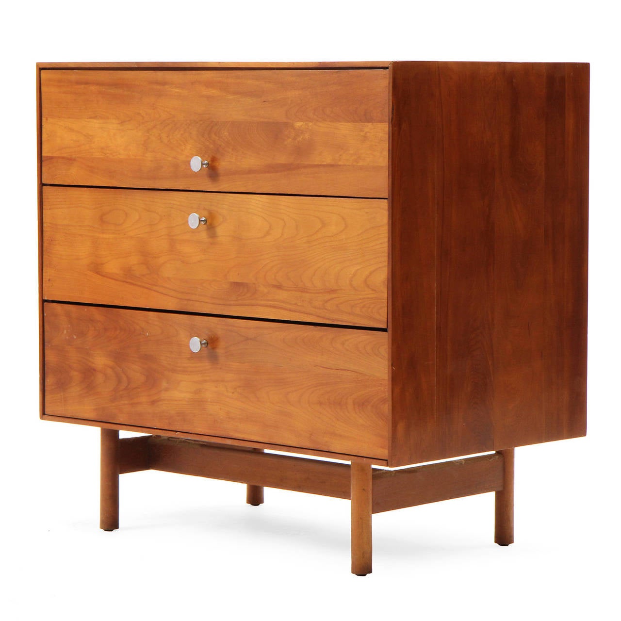A spare and refined chest of drawers having a thin-edged honey-toned birch rectilinear case with brushed steel drawer pulls floating on an architectural dowel leg base.