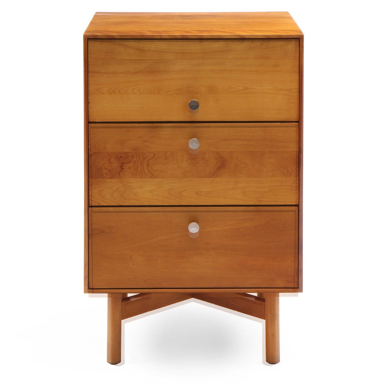 A narrow, spare and refined night stand having a thin-edged honey-toned birch rectilinear case with three drawers with brushed steel drawer pulls, floating on an x-braced architectural dowel leg base.