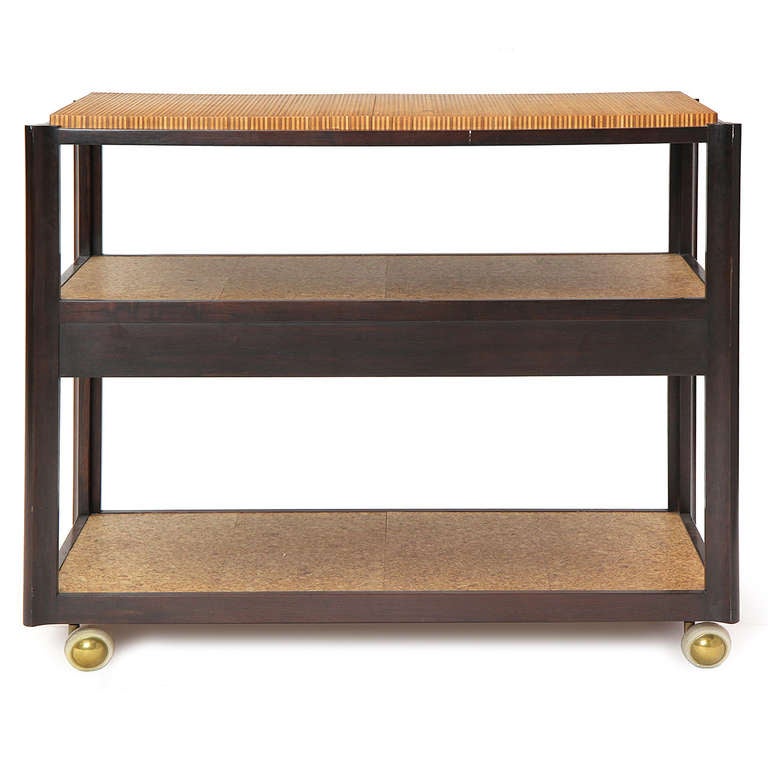 A two-tiered rectilinear bar cart made of walnut and cork having a floating central drawer and brass casters with a laminate end grain cutting board.