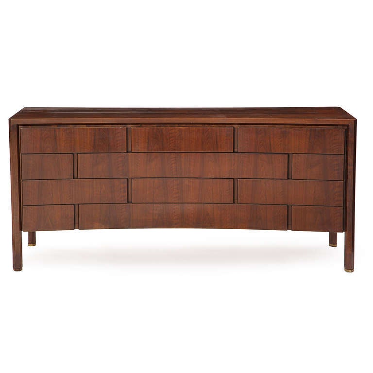 A stately and impressive bowed-front dresser with actively grain in dark walnut having twelve (12) staggered drawers and hexagonal legs with brass feet.