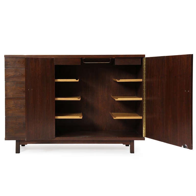 An asymmetrical wardrobe in solid walnut with exposed finger jointed joinery, having a shelved compartment accessed by bi-fold doors flanked by a bank of five drawers with exquisite brass hinged ring pulls, each with an inset of Natzler tile.