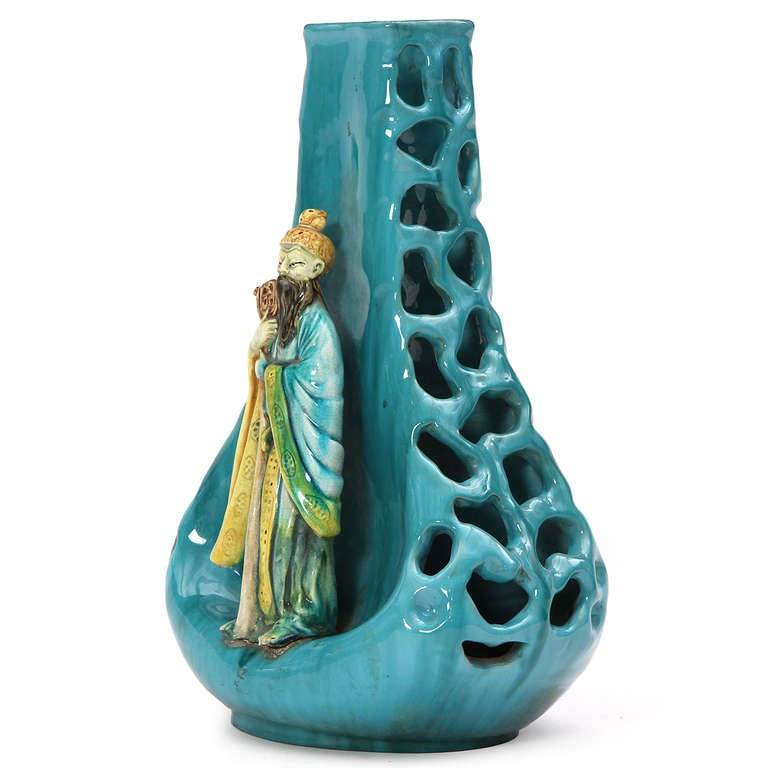A studio-made unique ceramic vase having a vividly poly chromed figure of a Chinese scholar standing perched on the edge of a dimpled and pierced base covered in a rich celadon glaze.