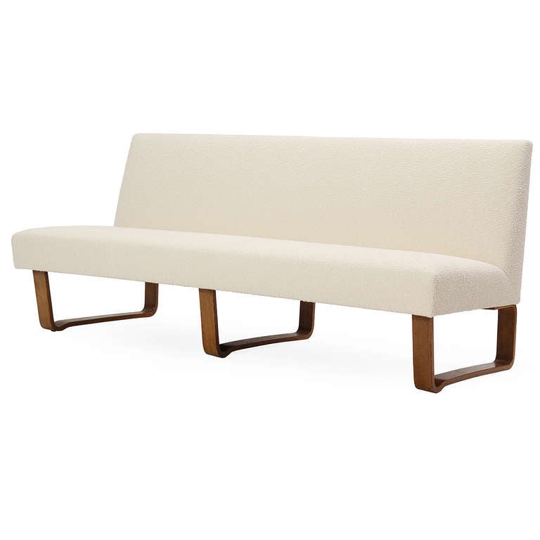 A striking and uncommon armless sofa having a beautifully-shaped seat in a rich cream upholstery floating atop three continuous laminated walnut 'sled' legs.