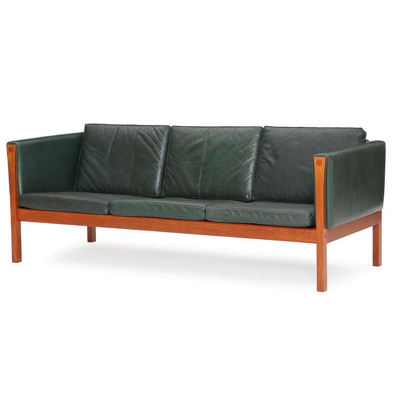 A striking even-arm sofa with slender dark emerald green leather covered cushions and an exposed teak frame.