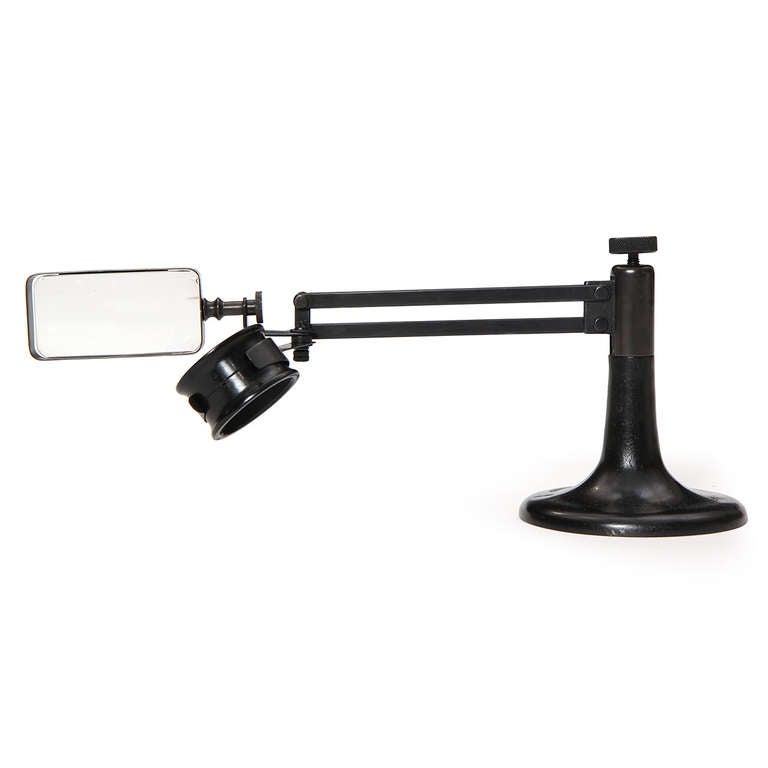 An industrial age magnifying glass and loupe on a cast iron pedestal base with an adjustable arm.