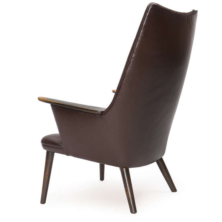 A high-back lounge with tight brown leather upholstery, exposed teak arms, and turned dowel legs.