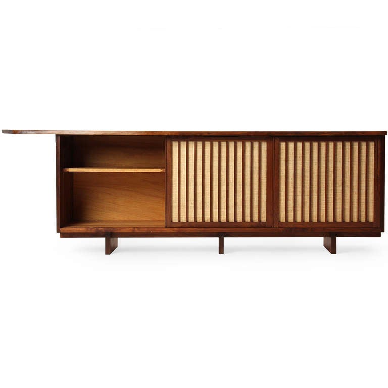 An excellent and long studio-made cabinet with a single board walnut top having a natural, free-edge overhanging top and three vertically slatted sliding doors (backed with pandanus cloth) which enclose a central bank of drawers flanked by shelved