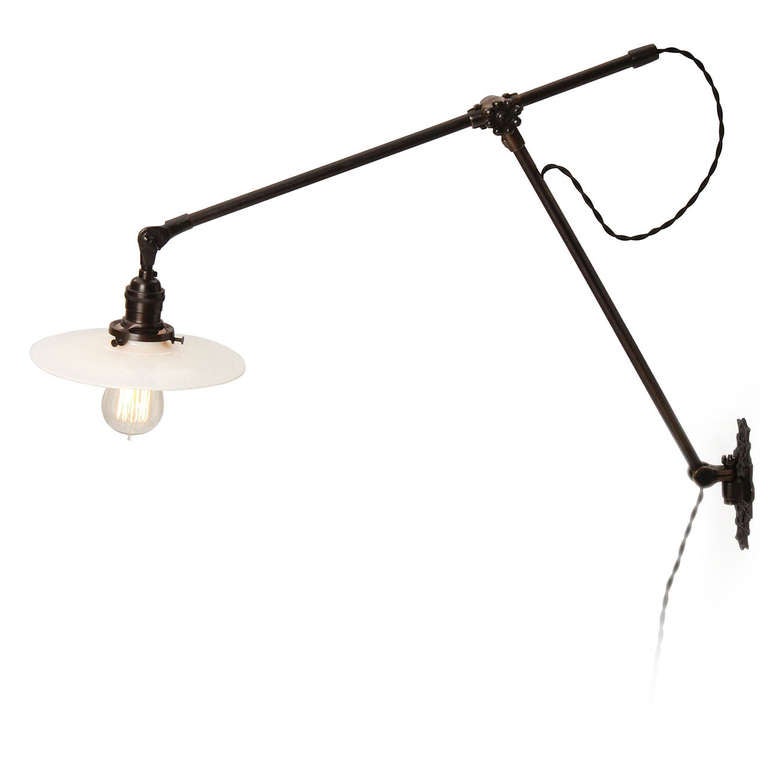 An early wall-mounted and fully articulated swing-arm lamp, having a dark patinated finish, milk glass disc shade and an ornate cast iron wall plate.