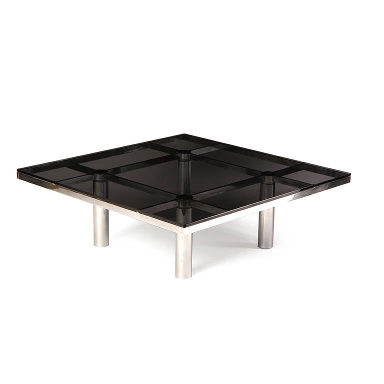 Une importante table basse carrée avec une lourde base en acier architectural grillagé supportant un épais plateau en verre fumé.