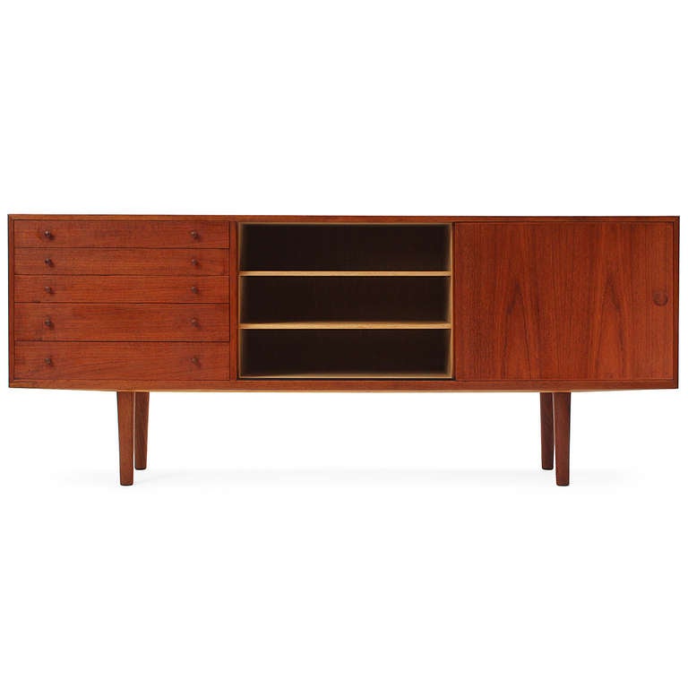 A credenza in warm teak with a bank of five drawers and twin sliding doors, floating on turned solid teak dowel legs.
