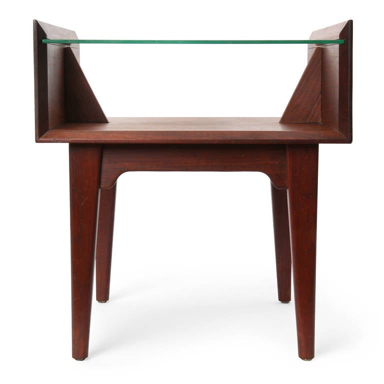 An interesting and finely crafted walnut end table having an architectural form with tapered legs and an expressive apron supporting a floating glass shelf.