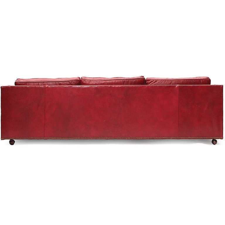 A very high quality and generously proportioned rectilinear three-seat sofa resting on six brass ball feet fully upholstered in rich polished red leather with down cushions, cylindrical bolster pillows and wonderfully accented with a continuous