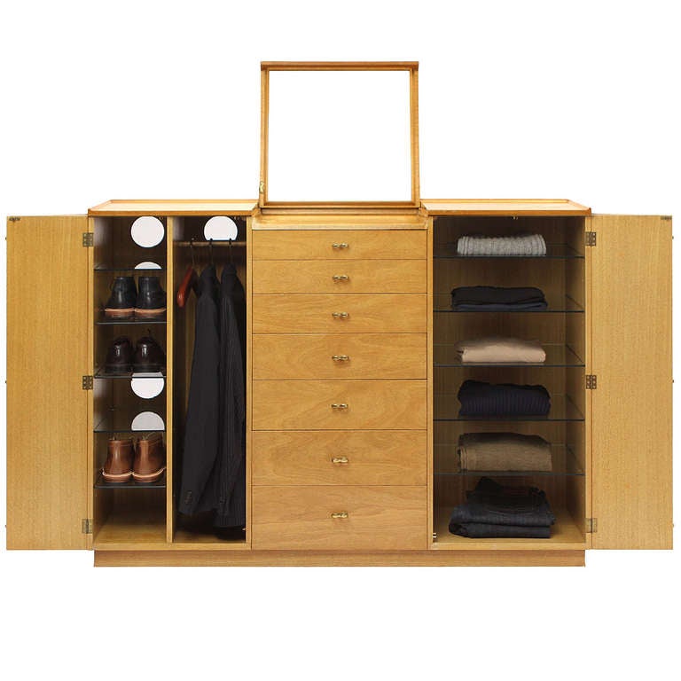 A masterful three-sectioned wardrobe in bleached mahogany. Having a central seven-bank of drawers and flanking closets-one with adjustable glass shelves, the other with shelving and a clothing rack. Additional details include a flip-up mirror and