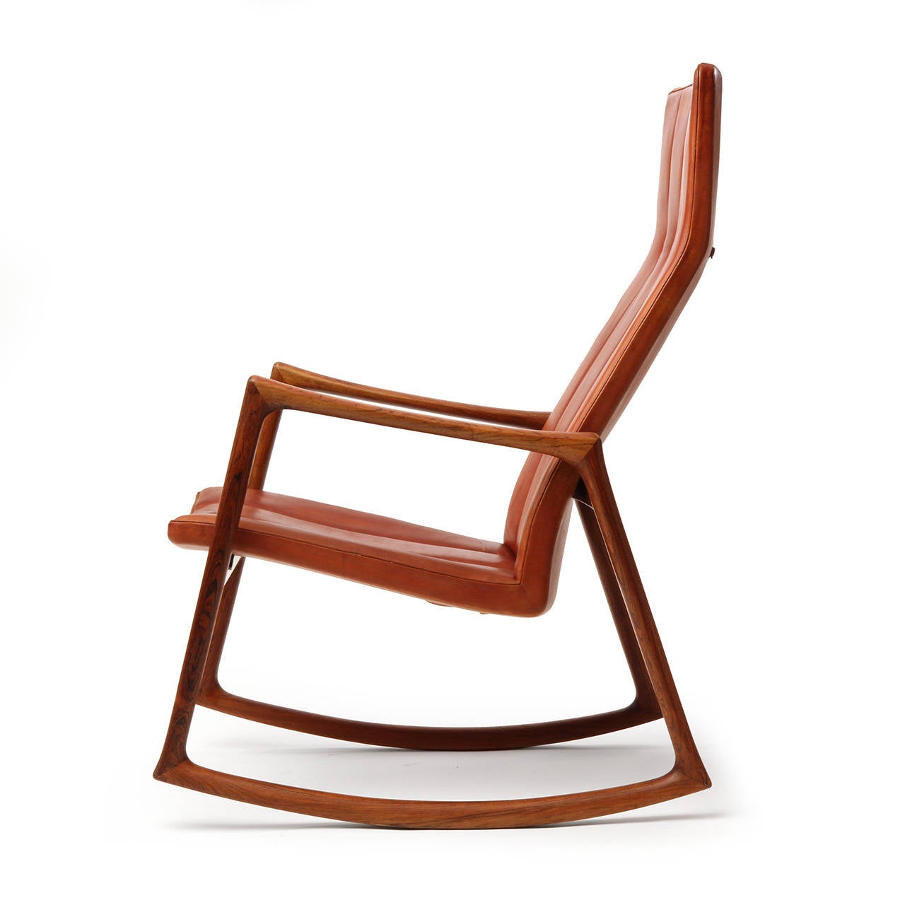 A sculptural, spare and superbly crafted rocking chair having an open frame of solid Brazilian rosewood supporting a floating ergonomic seat that retains its original tailored channeled caramel natural leather upholstery and adjustable headrest.