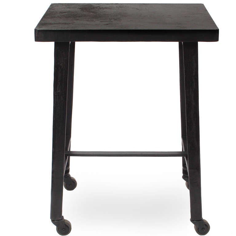 A tall industrial table having a forged steel square top and angled legs with casters and a flat steel 