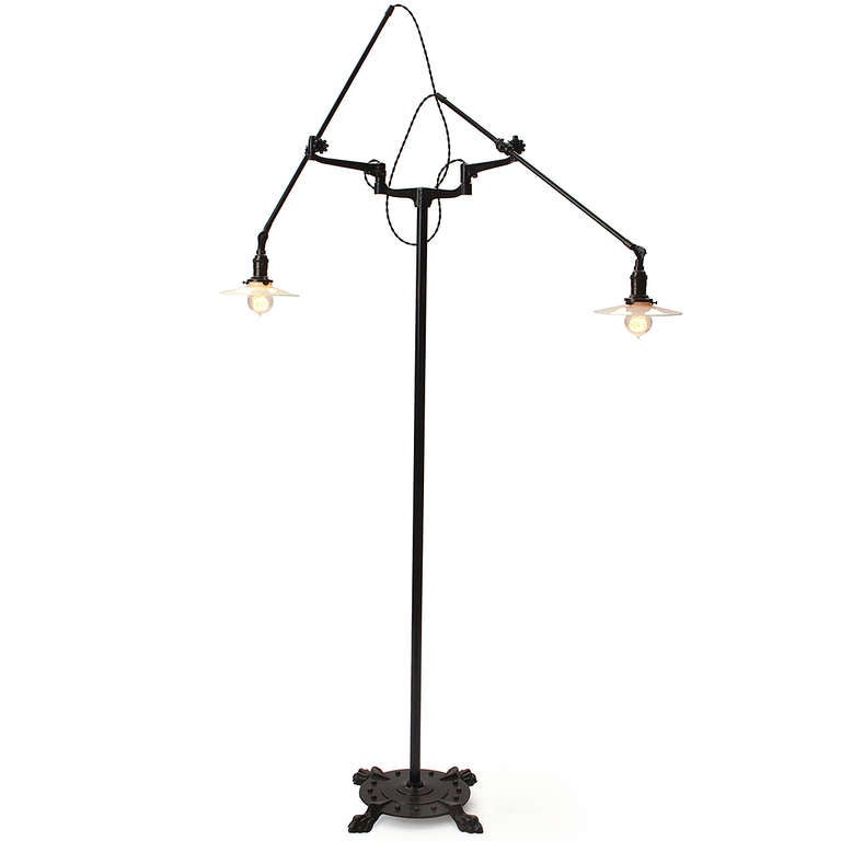 An industrial floor lamp in patinated steel and cast iron having a decorative footed base and two fully adjustable arms with milk glass shades. 