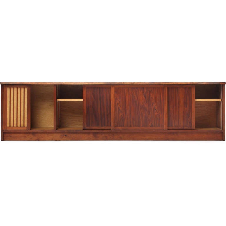 A fine and uncommonly long studio-made credenza by George Nakashima in highly figured black walnut having an expressive free-edge top, four sliding door panels (one panel with vertical slats and Pandus cloth) and exposed, hand-cut dovetail joinery.