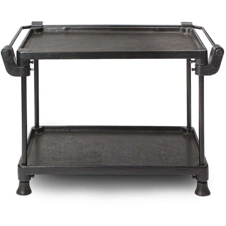 A heavy two-tiered industrial table in richly patinated steel and iron, having an external rail and shelves with raised edges, the entire form resting on distinctive bell-shaped feet.