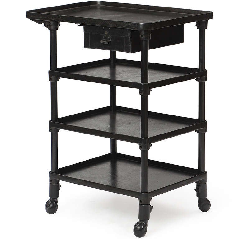 A tall four-tiered industrial table in patinated steel and iron, having adjustable shelves with raised edges and a floating drawer. The table rests on ball-and-claw feet.