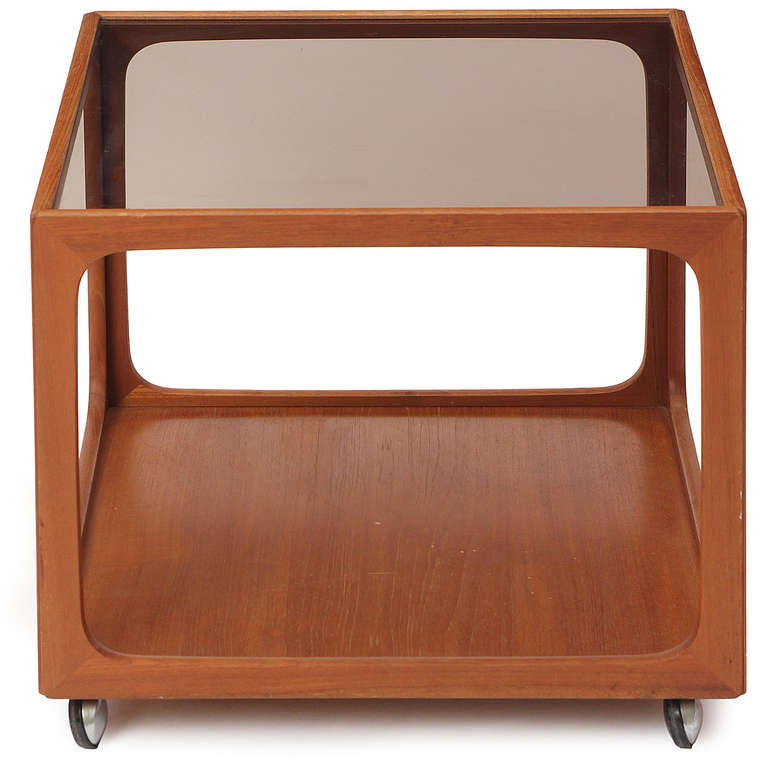 An open frame end table on casters in teak having a lower solid wood surface and floating glass top.