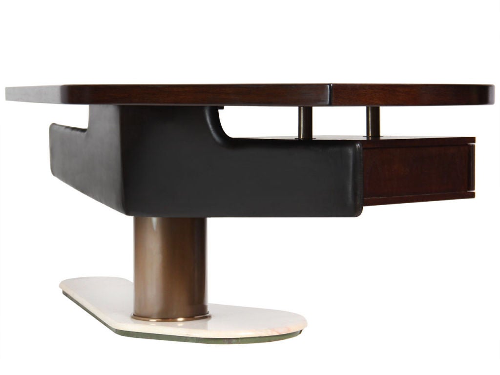 A unique asymmetrical desk with an ebonized walnut top and a padded leather modesty panel. Two floating drawers with leather fronts, and an off-center cylindrical bronze pedestal on a white marble base.