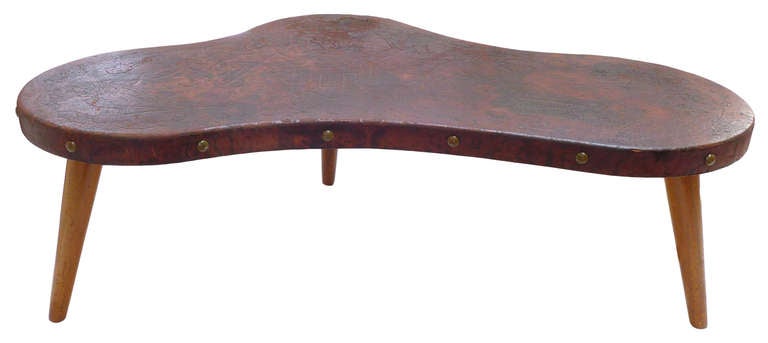 A wonderful, beautifully and exceptionally distressed, biomorphic coffee table clad in hand tooled leather. With its embossed pre-Columbian hunter-gatherer-themed surface and large brass studding, this piece is an interesting example of 1940s South
