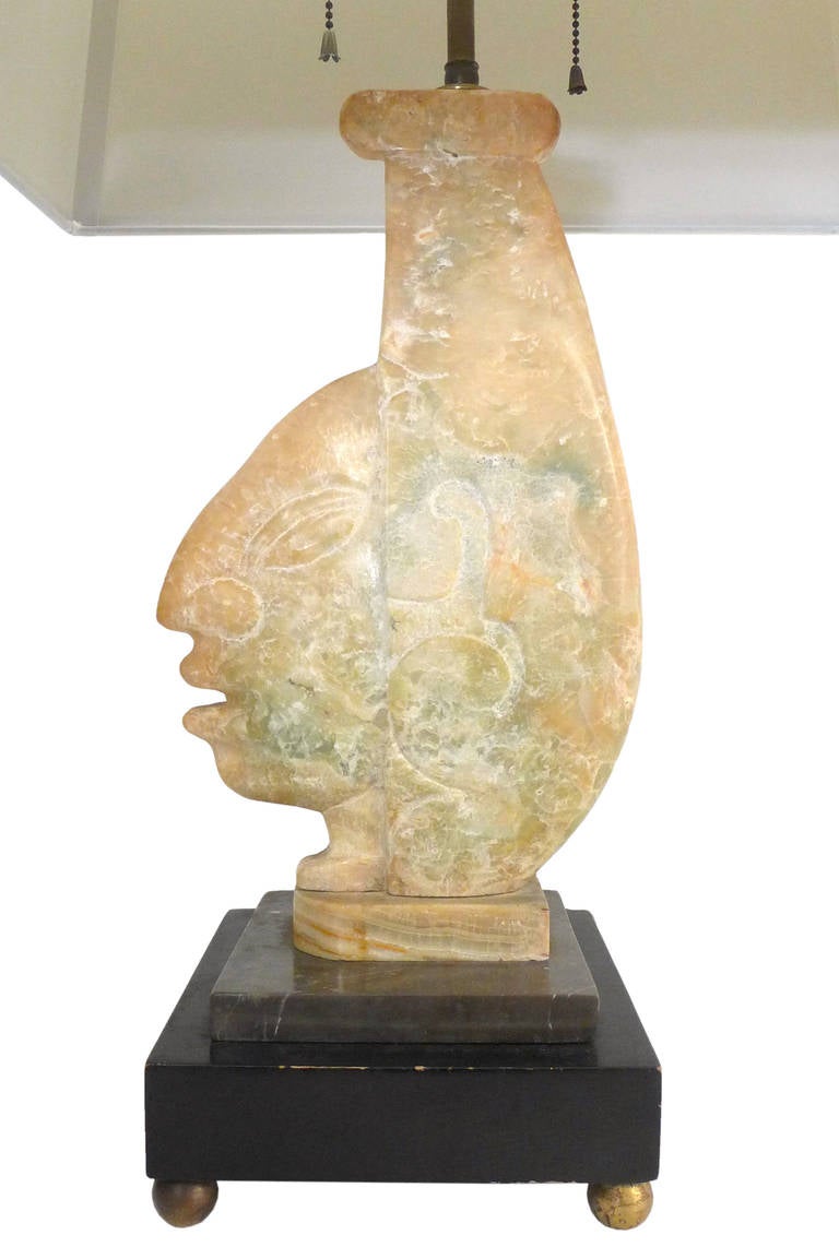 A wonderfully sculptural and powerful carved onyx lamp depicting a Pre-Columbian themed figure in profile. Primitive and modernist with beautifully executed carving on a lacquered and stepped wooden base with brass ball feet, matching spherical onyx