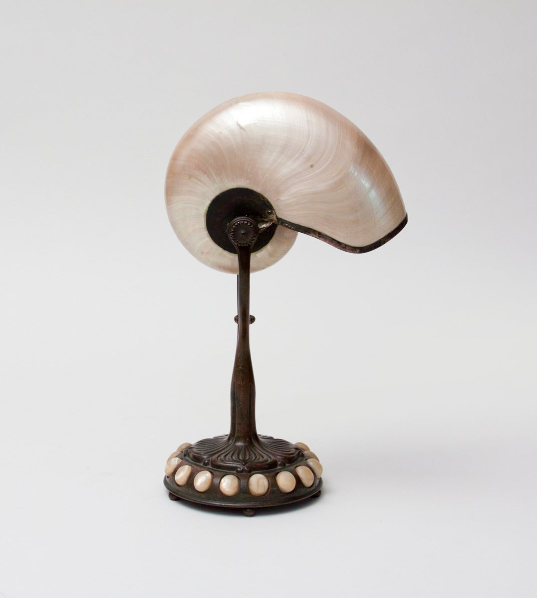 A Tiffany Studios desk lamp comprising an original nautilus shell shade on a bronze base, the leading and bronze finish in a rich brown patina. The base features an inset border of natural sea pebbles.