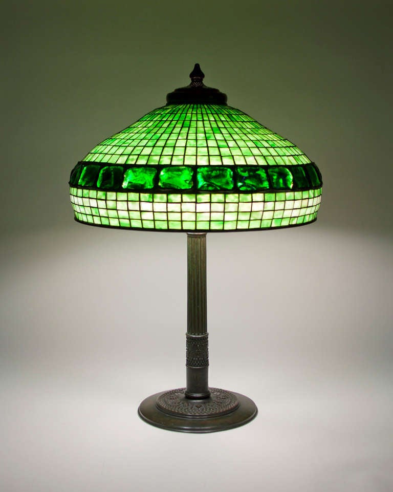 This Tiffany Studios Lamp comprises a geometric leaded glass shade in mottled green glass with an inset border of Tiffany Studios Favrile Glass Turtle Back tiles on a Roman Stick base. Louis C. Tiffany used the turtle back glass tiles throughout his