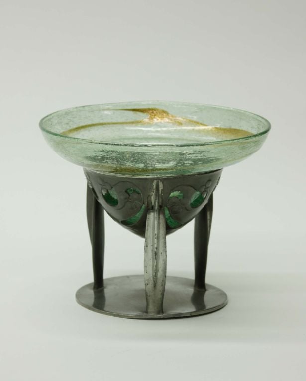 A three-legged Liberty and Co. pewter and glass fruit bowl, the stand designed by Archibald Knox and the clutha glass liner by Christopher Dresser.  The glass in a translucent green with aventurine trailings.