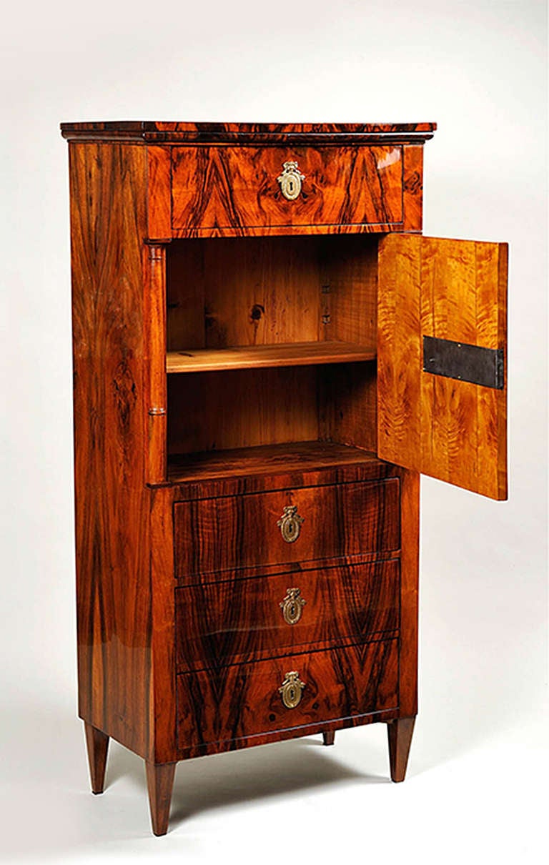 A tall Biedermeier cabinet with book-matched walnut veneer, four drawers with center cupboard and one adjustable shelf concealed by a door. Tapered legs, hand rubbed shellac polish.