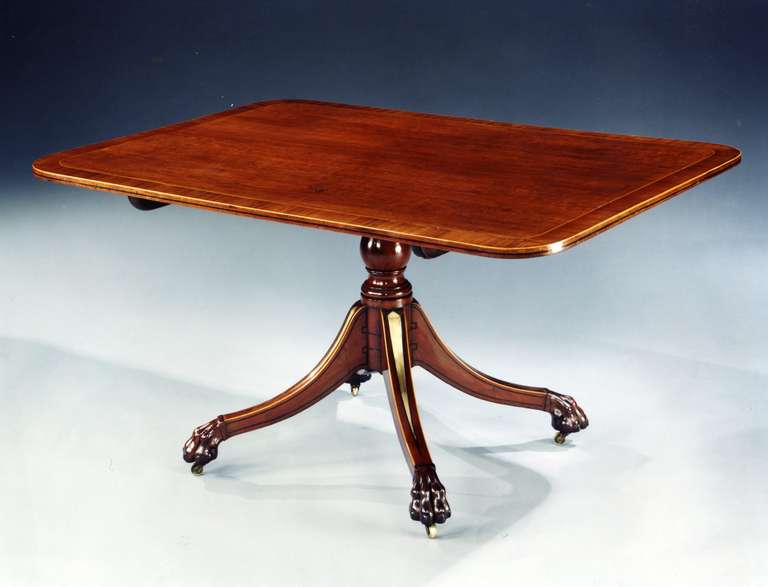 An early 19th century plum pudding mahogany brass inlaid and ebony strung breakfast table; the rounded rectangular molded tilt-top with mahogany crossbanding, on a baluster stem on quadripartite base with spay legs inlaid with tapering brass panels