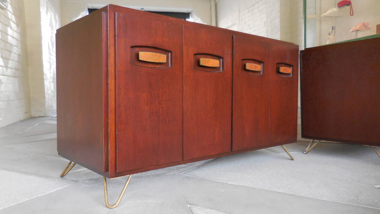 A dynamic pair of solid mahogany low cabinets from the 1950s with elongated enameled brass door pulls. This pair was acquired from a home in Palm Springs along with several other pieces, including a three-drawer chest and a six-drawer chest which