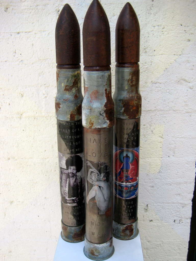 A powerfully provocative sculptural trio of wooden and steel World War II dummy artillery shells emblazoned with non-violent credo and imagery. Made in Brooklyn for use by the Navy from 1942- 1944 these weigh about 22 lbs. each. The first shell has