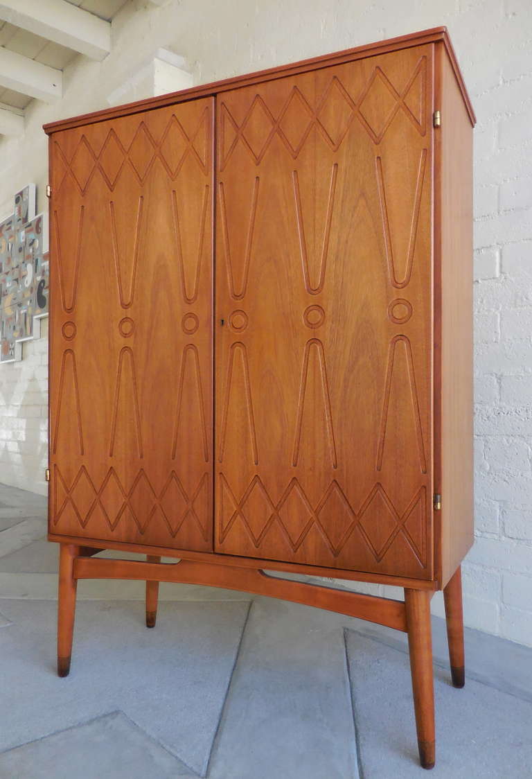 A sophisticated Swedish mahogany cabinet on legs from the 1960's.  The upper structure of the piece has graphically patterned incised doors and the cabinet sits up on splayed legs with brass cup sabots.  The interior has one stationary and two