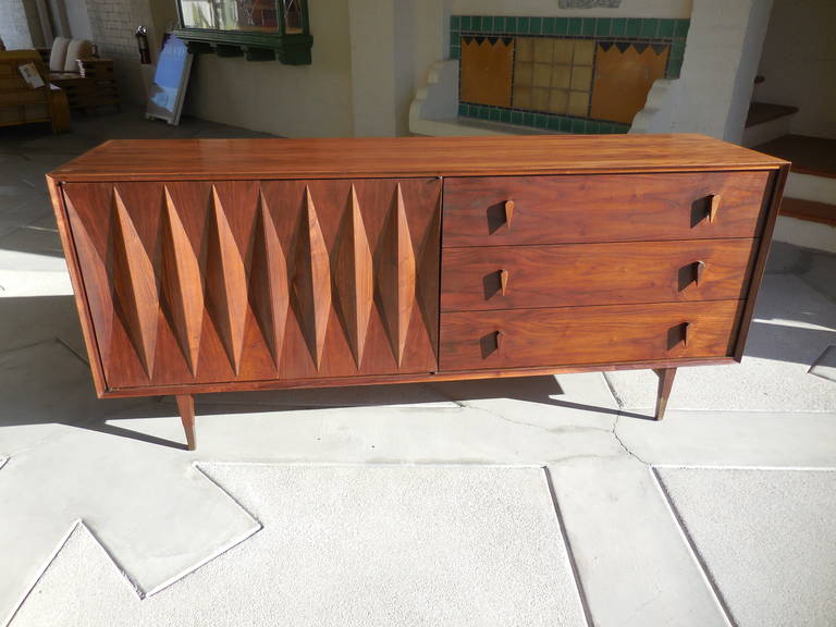 A sculptural walnut credenza or cabinet, designed in the 1950s by interior designer Albert Parvin. The credenza is divided into two segments; on the left side a pair of deeply sculpted doors with diamond-shaped details open to reveal three sliders,
