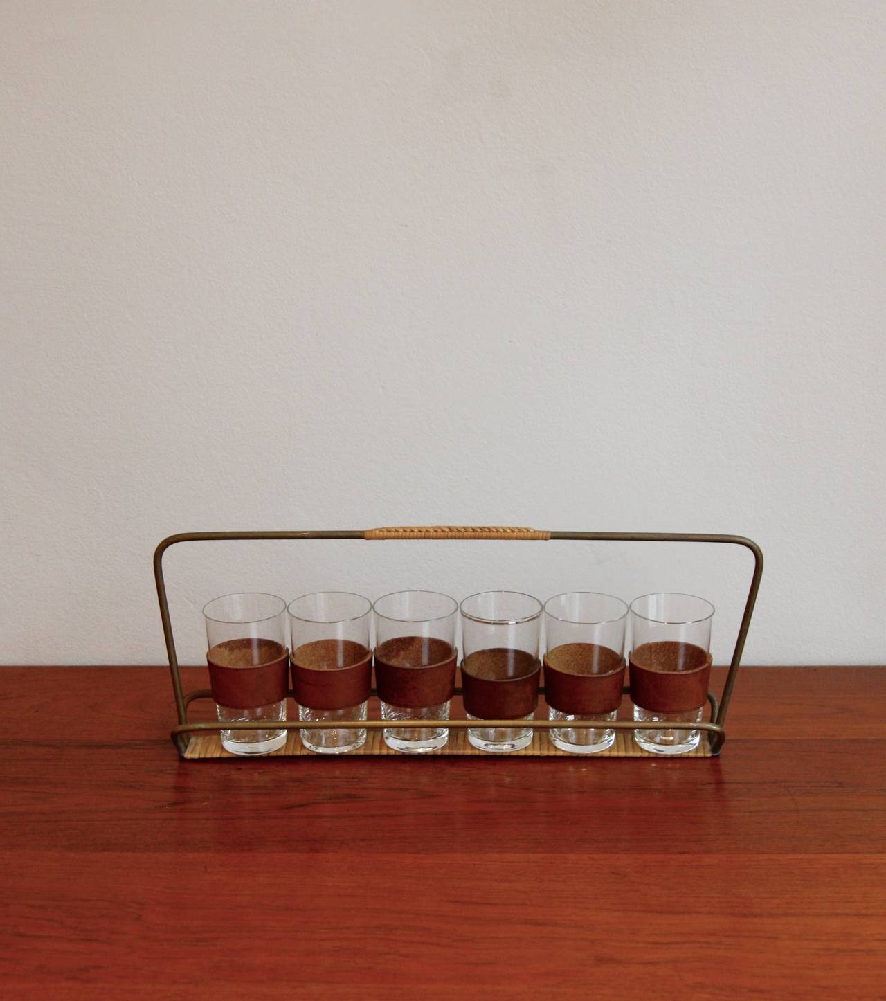 Set of six small glasses with cognac leather cover and brass and wicker drinks carrier handmade in the workshop of Carl Auböck's in the 1950s. Glasses are 10 cm in height and 5.5 in diameter while the carrier is 39.5 cm long, 14 cm tall and 6 deep.