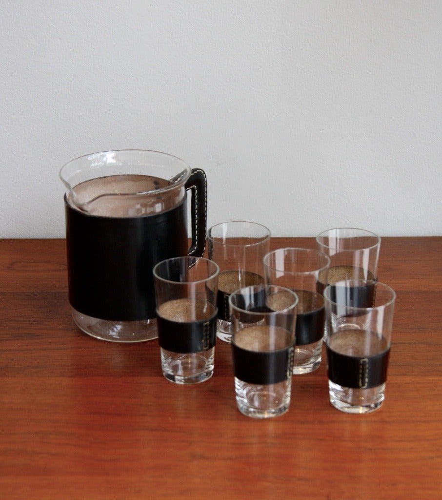The pitcher, measuring 15 cm in height and 11 in diameter, together with the glasses - 10 and 5.5 cm - constitute an elegant set of seven handmade glasses pieces covered in black leather. Made in the Viennese Carl Auböck's Werkstatte in the 1950s.