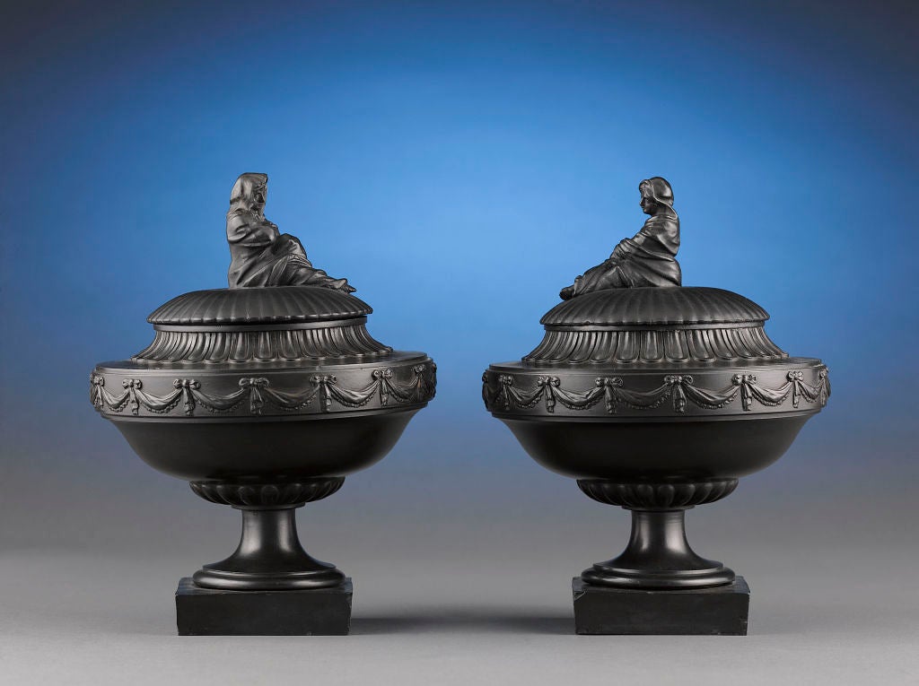This remarkable pair of black basalt covered urns was crafted by the inimitable Wedgwood & Bentley. The Neoclassical design incorporates architectural detailing such as swags, egg and dart accents and two allegorical figures representing Winter and