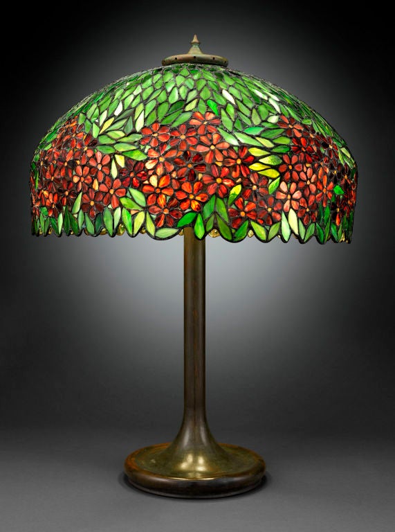 This monumental Handel lamp boasts a stunning stained leaded glass shade, its pattern displaying bright red flowers in a field of verdant green leaves. Set atop a deeply patinated bronze base, this lovely Art Nouveau lamp boasts excellent