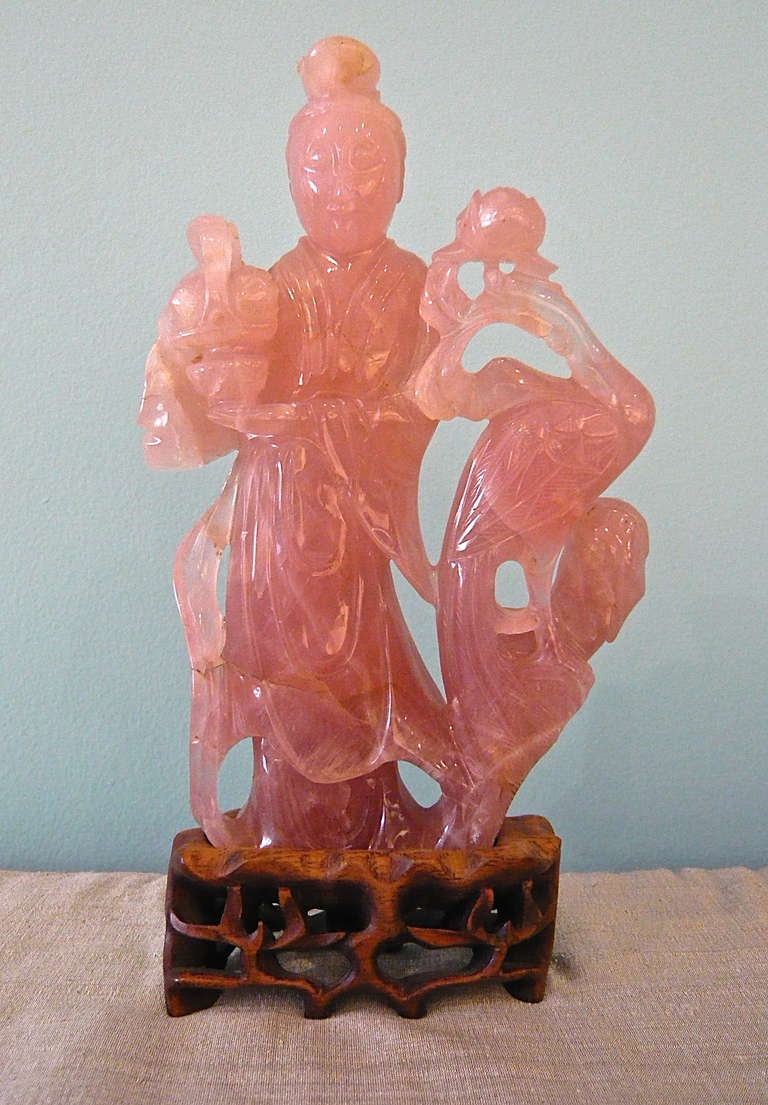 A beautiful and elegant rose quartz figure of a deity in flowing robes holding a basket of fruit in one hand with a crane and lotus flower on a wooden stand.