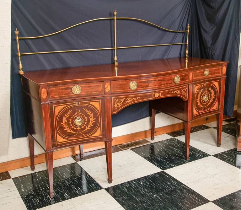 English, inlaid mahogany serpentine front sideboard with arched brass rail. Intricate satinwood, olivewood and ebony wood inlays decorate the front of the elegantly proportioned sideboard. A large, central silver drawer is flanked by a smaller