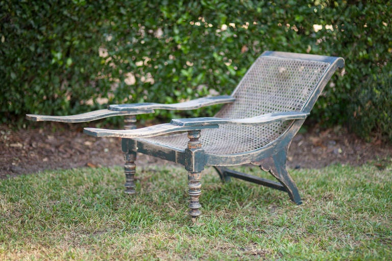 A mid-20th century planter’s chair in the British Colonial style; with extending boot rests, turned front legs, gently worn distressed paint of a gun-metal gray and black with the wood showing through in places. Caning in excellent condition; the